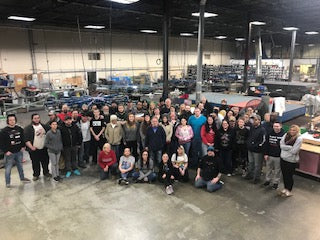 A large group of people posing for a group photo inside a spacious industrial workshop or factory with machinery and metal structures in the background.